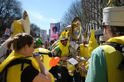 A spirited brass ensemble clad in vibrant yellow ponchos performs amid a political demonstration in Portland's historic Old Town district. The tubist at center, wearing sunglasses and a broad grin, anchors the musical protest as fellow musicians brandish their instruments like banners of resistance. Behind them, protest signs punctuate the crowd while bare winter trees frame the scene against a crisp blue sky, creating a striking tableau of civic engagement through music.