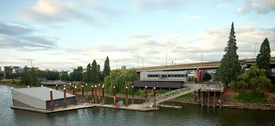 Portland Fire & Rescue Station 21 sits anchored on the Willamette River beneath the gentle light of an overcast evening sky. The modern floating firehouse, distinguished by its corrugated metal roof and red support pilings, connects to shore via a steel gangway, creating a unique waterfront emergency response facility. Towering Lombardy poplars frame the scene while the Hawthorne Bridge spans the background, establishing the station's strategic location in Portland's Central Eastside industrial district.