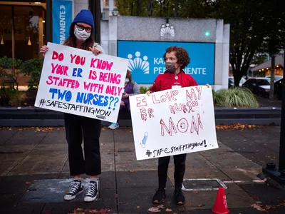 A woman in a cobalt blue knit beanie and face mask holds a handwritten protest sign questioning emergency room staffing levels, while a young child beside her displays a heartfelt message of support for their nurse mother. The pair stands on autumn-scattered sidewalks outside Kaiser Permanente's towering headquarters in Portland's Lloyd District, their white poster boards catching the soft urban light against the healthcare giant's signature blue corporate signage. The intimate scale of their grassroots demonstration contrasts powerfully with the institutional architecture looming behind them.