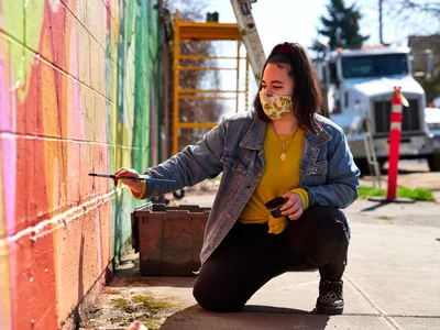 Multidisciplinary artist Vilatose kneels on the sidewalk, paintbrush in hand, adding vibrant details to 'Jardín Cubana,' a colorful mural depicting three female figures along a brick wall in Portland's Alberta Arts District. Bathed in warm afternoon sunlight, she wears a yellow knit sweater beneath a denim jacket and a patterned face mask, her dark hair pulled back as she focuses intently on her work. Behind her, construction equipment and urban infrastructure frame this moment of artistic creation in the heart of Northeast Portland's creative corridor.