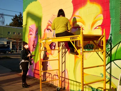 Artist Villatose works from a yellow scissor lift to paint 'Jardín Cubana,' a vibrant mural depicting three female figures on a brick wall along Northeast Alberta Street in Portland's Alberta Arts District. A masked collaborator documents the process from below, capturing the creative energy of this urban art installation. The mural features bold yellows, pinks, and greens that transform the building's facade, while late afternoon light creates dramatic shadows across the industrial equipment and colorful artwork.