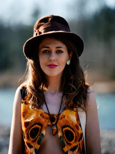 A young woman with flowing brunette hair poses confidently along the Floodplain Trail riverbed in Multnomah County, Oregon, her striking butterfly-wing printed top catching the warm afternoon light. Her wide-brimmed felt hat casts gentle shadows across her face while bold pink lipstick and delicate pearl earrings add vintage elegance to her bohemian style. The soft bokeh background of Pacific Northwest riverbed landscape creates an dreamy, ethereal atmosphere that highlights her direct gaze and natural beauty.