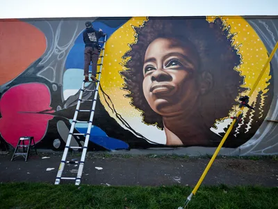 Artist Travis Fields, known as campographic, stands atop an aluminum ladder while painting a vibrant mural of poet Amanda Gorman on a wall in Northeast Portland's Alberta district. The luminous portrait depicts Gorman with her natural hair flowing against a golden celestial backdrop dotted with stars, her gentle smile radiating hope and determination. The surrounding composition features bold geometric shapes in coral, magenta, and azure blue, creating a dynamic frame around the central figure. Paint supplies and equipment scattered on the weathered pavement below speak to the ongoing creative process transforming this urban canvas.