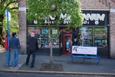Two men pause on the sidewalk outside Music Millennium, a beloved Portland record store on East Burnside Street, their casual stance suggesting the unhurried rhythm of neighborhood browsing. Hand-painted window advertisements for Record Store Day and upcoming performances create a vibrant collage against the brick storefront, while dappled sunlight filters through spring foliage overhead. The scene captures the enduring appeal of independent music retail in Portland's eclectic Kerns neighborhood, where vinyl culture thrives amid the city's creative community.