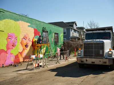 Multidisciplinary artist Villatose works from scaffolding to paint a vibrant pop art mural titled 'Jardín Cubana' featuring three stylized female figures on a green cinder block wall along Northeast Alberta Street in Portland's Alberta Arts District. The scene captures the intersection of urban art and community development, with residential houses visible in the background and construction equipment stationed nearby. Bright afternoon light illuminates the bold pink, yellow, and red palette of the emerging artwork, while safety barriers and a white commercial truck frame this moment of creative transformation in the gentrifying neighborhood.