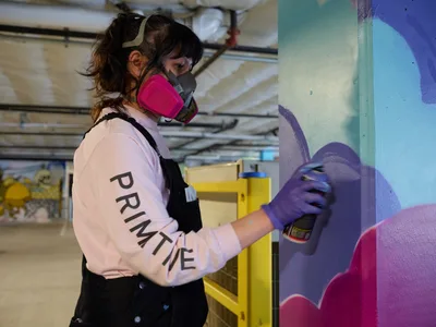 A focused muralist wearing a pink respirator mask and "PRIMITIVE" sweatshirt applies purple spray paint to a vibrant wall mural in the converted parking garage of Clay Creative in Southeast Portland's Hosford-Abernethy neighborhood. The industrial workspace, with its exposed ceiling infrastructure and yellow storage units, provides an authentic urban canvas for the emerging artwork's bold blues, purples, and magentas.