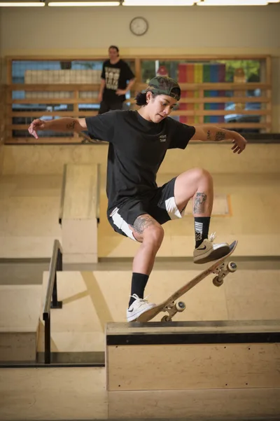 Pro skateboarder Christiana Means executes a precise frontside carve across the curved concrete of Stronger Skatepark in Milwaukie, Oregon. Her tattooed arms extended for balance, she navigates the bowl's transition with fluid control while fluorescent lights cast an even glow across the indoor facility. The geometric lines of the skate park's wooden ramps create a backdrop of angles and shadows, with another skater visible in the distance.