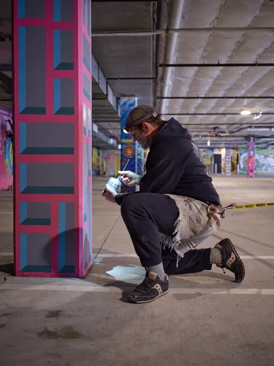 Portland artist and bike activist Rabbitt kneels in focused concentration within the Clay Creative building's parking garage, spray painting his signature geometric patterns onto a concrete column. The harsh fluorescent lighting casts dramatic shadows across the industrial space, illuminating vibrant pink and blue geometric forms that transform the utilitarian concrete into urban canvas. His weathered work clothes and protective gear speak to the physical dedication required for large-scale mural creation in Portland's creative underground.