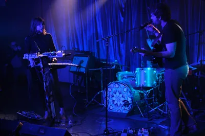 Experimental band Sun Blood Stories, originally from Boise, Idaho and now based in Portland, performs in the intimate confines of Dante's venue during Rx Fest, bathed in deep cobalt stage lighting that transforms the space into an ethereal sonic cathedral. The keyboardist, silhouetted against flowing curtains, commands an array of synthesizers while the drummer maintains rhythm behind a kit adorned with geometric mandala artwork. Cables snake across the stage floor as purple and blue washes create dramatic shadows, capturing the mesmerizing live sound that has been likened to modern-day opera.