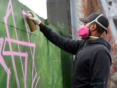 A dedicated muralist wearing a bright pink respirator mask and gray hoodie applies fresh pink paint to a vibrant green wall in the parking lot of Clay Creative in Portland's Hosford-Abernethy neighborhood. The artist's focused concentration is evident as he carefully guides the spray can across the concrete surface, adding geometric lines to what appears to be an emerging street art piece. The industrial setting and protective gear speak to the serious craft of contemporary urban art creation.