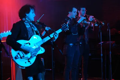 Charlotte McCaslin of Roselit Bone commands the stage with her hollow-body electric guitar, bathed in saturated cyan and crimson stage lighting at Dante's in Portland. The intimate venue's moody atmosphere envelops the trio as brass players behind her contribute to the sonic landscape. The dramatic color temperature creates an otherworldly glow against the dark venue backdrop, capturing the raw energy of Portland's underground music scene.