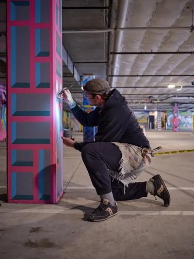 Portland bike activist and artist Rabbitt crouches in concentrated focus, applying precise brushstrokes to a vibrant geometric mural on a concrete pillar. The industrial parking garage of Clay Creative building provides a stark backdrop, its exposed ceiling and fluorescent lighting contrasting with the artist's colorful pink, blue, and gray geometric patterns. Paint cans rest nearby as Rabbitt, wearing a knit cap and dark hoodie, transforms the utilitarian space into an urban canvas.