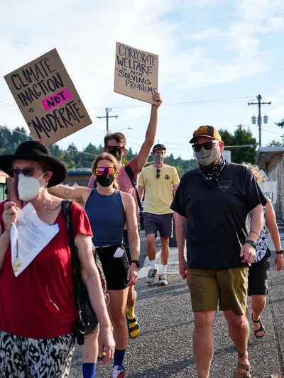 Masked protesters stride purposefully down a sun-dappled street in Oregon City, Oregon, their handmade signs demanding climate action and critiquing corporate welfare. The diverse group of activists moves with quiet determination beneath a partly cloudy sky, their colorful summer clothing contrasting against the stark black lettering of their protest signs. Power lines stretch overhead while residential buildings frame this grassroots demonstration for environmental justice.