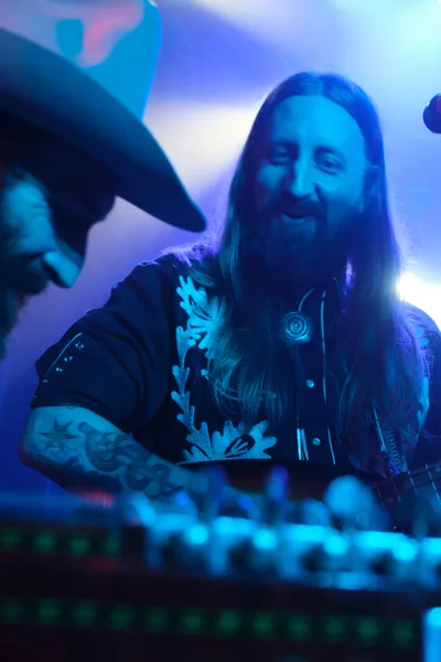 A bearded musician with flowing hair grins widely while performing behind an electronic mixing console, bathed in vibrant blue and purple stage lighting. The intimate venue atmosphere pulses with energy as the artist, part of electronic duo Roselit Bone, manipulates controls during their RX Fest 2019 performance. Tattoos visible on his arms catch the atmospheric lighting while the background dissolves into a dreamy wash of concert illumination.