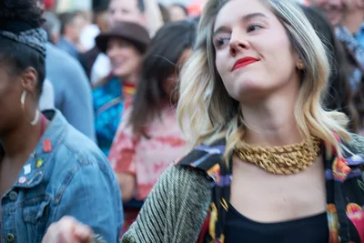 A young blonde woman with crimson lips and a statement gold chain necklace gazes upward with serene satisfaction, captured in the warm glow of late afternoon light at White Owl Social Club's summer day party. The shallow depth of field blurs the denim-clad crowd behind her into soft bokeh, creating an intimate portrait against the bustling Portland social scene. Her metallic chain catches the golden light while partygoers fade into impressionistic shapes of blue and coral hues.
