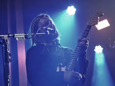 Russell Marsden of Band of Skulls delivers an electrifying performance at Webster Hall during the band's 10 Year Anniversary Tour. Bathed in ethereal blue and cyan stage lighting, the guitarist-vocalist leans into his microphone while gripping his electric guitar, his shoulder-length hair catching the atmospheric glow. The intimate venue's lighting creates a dreamy haze around the performer, capturing the raw energy and nostalgic celebration of a decade-long musical journey in Manhattan's historic music district.