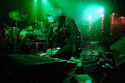 Electronic musician Eli Savage, performing as Ellipsism, hunches intently over his mixing console at RXFest 2019, bathed in ethereal green stage lighting that transforms Dante's intimate venue into a sonic laboratory. His silver hair catches the emerald glow as he manipulates analog controls, surrounded by a constellation of synthesizers, drum machines, and electronic equipment that sprawl across the darkened stage. The atmospheric lighting creates dramatic shadows and highlights, emphasizing the concentrated focus of the artist as he sculpts soundscapes in Portland's legendary underground music venue.