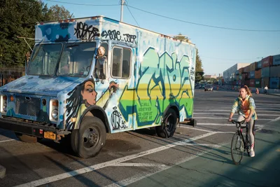 Golden hour light bathes a vibrant graffiti-covered food truck parked along Kent Avenue in Brooklyn's Navy Yard district, its weathered white surface transformed by bold blues, yellows, and greens depicting stylized figures and tags. A young woman on a bicycle pauses beside the mobile canvas, her figure casting long shadows across the asphalt as industrial shipping containers stack in the soft-focused background. The scene captures the raw creative energy of Brooklyn's waterfront, where street art culture intersects with everyday urban life.