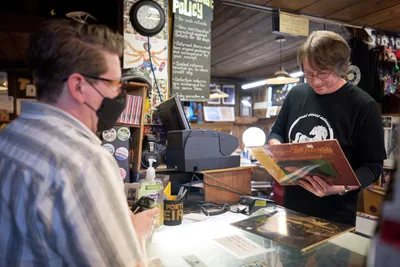 Inside the iconic Music Millennium record store on East Burnside in Portland's Kerns neighborhood during Record Store Day 2022, a bespectacled shopkeeper in a black t-shirt carefully examines a vinyl album while a masked customer waits at the counter. The intimate scene unfolds beneath warm pendant lighting, surrounded by towering shelves of records and memorabilia. A vintage wall clock and handwritten store policy sign create an authentic atmosphere of analog music culture, while the glass counter displays rare finds below.