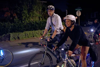 Two helmeted cyclists pause during Portland's Drop Out Prom Ride, part of the city's annual Pedalpalooza festival in June 2019. The man in a white dress shirt and tie contrasts with the woman in dark attire as they navigate the nighttime streets of Southeast César E. Chávez Boulevard. LED bicycle lights pierce the darkness while lush greenery creates a natural backdrop, capturing the playful intersection of formal wear and urban cycling culture during this unconventional prom celebration.
