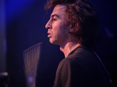 Julian Dorio, drummer for Band of Skulls, captured in profile during the band's 10 Year Anniversary Tour at Webster Hall in Manhattan. Theatrical blue and purple stage lighting sculpts his concentrated expression as he gazes into the distance, his tousled hair catching the ambient glow. The intimate venue's atmosphere envelops the scene in deep shadows, with the suggestion of his drum kit visible in the soft focus background.