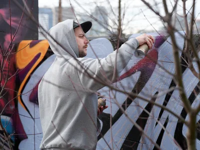 Travis Fields, known as Campographic, concentrates on his craft as he applies spray paint to a weathered concrete wall at the Clay Creative Building in southeast Portland's Hosford-Abernethy neighborhood. Bare winter branches create a natural frame in the foreground, while his grey hoodie and baseball cap provide stark contrast against the emerging purple and red hues of his urban artwork. The overcast Pacific Northwest light filters through the scene, capturing this moment of artistic creation in Portland's vibrant street art community.