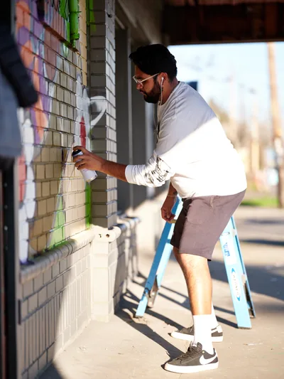 Artist Heysus concentrates intently as he applies spray paint to a cream-colored brick wall, working from a blue stepladder on a sun-drenched sidewalk in Northeast Portland. The warm afternoon light illuminates his focused expression and white long-sleeved shirt as he adds vibrant details to his emerging mural. Green tape marks his careful preparation work against the textured brick surface, while the soft-focus urban backdrop hints at the Alberta Arts District's creative energy.