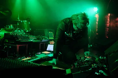 Electronic musician Eli Savage, performing as ellipsism, becomes fully absorbed in his craft at RXFest 2019, hunched over an intricate web of synthesizers and mixing equipment at Portland's intimate Dante's venue. Ethereal green stage lighting bathes the performance space, creating an otherworldly atmosphere that mirrors the experimental nature of his electronic compositions. His flowing hair catches the colored light as he manipulates controls with intense concentration, embodying the solitary focus required for live electronic music creation.