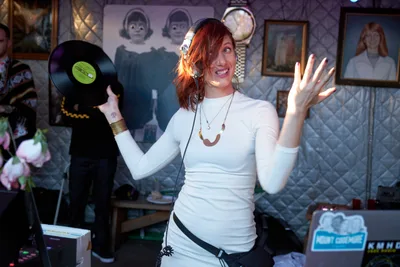 DJ Doctor Dakar commands the dance floor with infectious energy at White Owl Social Club, her auburn hair catching golden light as she holds a vinyl record aloft while gesturing enthusiastically to the crowd. The intimate Portland venue's eclectic wall art creates a bohemian backdrop of vintage portraits and quilted textures. Her white fitted top and layered jewelry embody the relaxed sophistication of Sunday afternoon party culture in the Pacific Northwest.