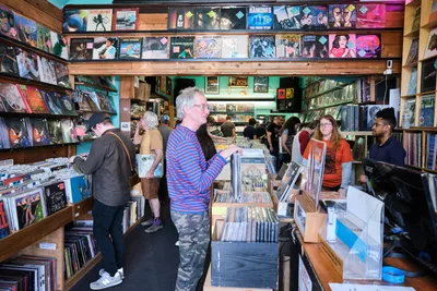 Inside the beloved Music Millennium on East Burnside, Portland's iconic record store pulses with afternoon energy as vinyl enthusiasts navigate densely packed aisles. A silver-haired collector in a blue striped shirt examines albums near towering wooden bins, while other customers browse the extensive collection that climbs floor-to-ceiling along warm wood shelving. The shop's mint green walls and eclectic atmosphere create an intimate sanctuary for music discovery.