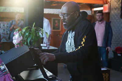 DJ Cee White commands the turntables at White Owl Social Club during Your Sunday Best day party, his weathered hands working the equipment with practiced precision. Golden afternoon light streams through the Portland venue, illuminating his focused expression behind wire-rimmed glasses as patrons gather in the warm, intimate space. Fresh flowers accent the DJ booth while the eclectic club atmosphere buzzes with anticipation.