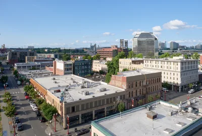 An elevated perspective captures Portland's Historic Chinatown district on a brilliant summer afternoon, where century-old brick buildings with ornate facades stand alongside modern glass towers. The warm golden light bathes the mixed-use streetscape, revealing the neighborhood's layered architectural evolution from historic commercial blocks to contemporary urban development. Tree-lined streets create geometric patterns between the vintage storefronts and newer residential buildings, while the distinctive domed tower of a prominent downtown building punctuates the distant skyline.