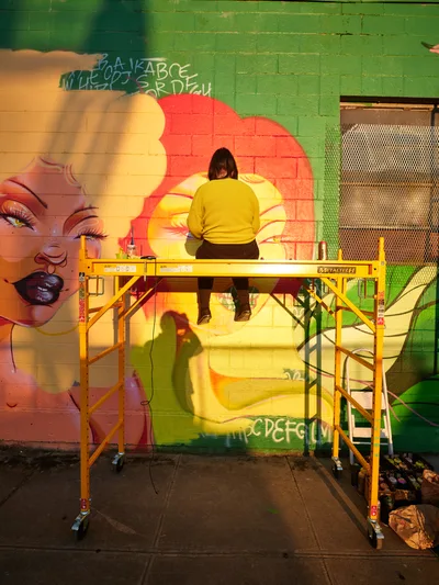 Multidisciplinary artist Vilatose stands on yellow scaffolding, painting the radiant mural 'Jardín Cubana' featuring three female figures on Alberta Street in Portland's arts district. The warm golden light bathes the scene as the artist works in a bright yellow sweater, adding finishing touches to the colorful pop art piece that transforms the urban brick wall. Paint supplies and brushes are scattered on the scaffolding platform below, while the vivid oranges, pinks, and greens of the artwork create a striking contrast against the emerald brick facade.