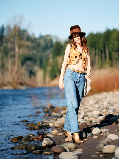 A young woman in bohemian attire stands confidently on a pebbled riverbank along Oregon's Floodplain Trail. Golden hour light illuminates her vintage-inspired ensemble of flared jeans, graphic tank top, and distinctive wide-brimmed hat as she poses beside gently lapping waters. The shallow river meanders through a backdrop of towering evergreens and deciduous trees, creating an intimate wilderness portrait that captures both personal style and natural serenity.