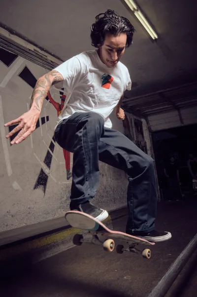 A tattooed skateboarder in a white graphic tee executes a fluid movement within the weathered concrete bowl of Commonwealth Skateboarding in Portland's Buckman neighborhood. Harsh fluorescent lighting cuts through the subterranean atmosphere, casting dramatic shadows across the curved transitions and worn surfaces. The skater's focused expression and extended arm suggest a moment of balance and control as his board glides through the empty pool, embodying the raw authenticity of underground skate culture.