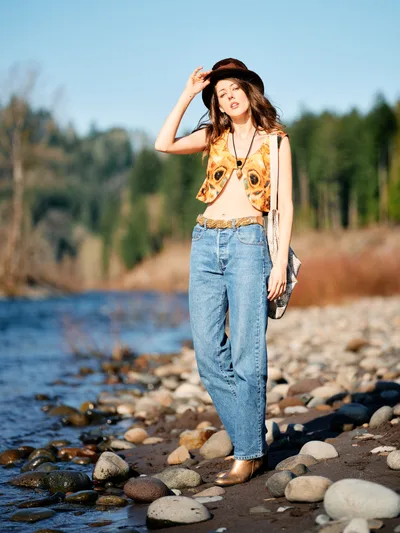 A young woman in bohemian attire poses confidently along the rocky shoreline of the Floodplain Trail in Multnomah County, Oregon. She wears a vibrant butterfly-print vest over a cream top, high-waisted denim, and a dark fedora while touching the brim with one hand. The golden hour light illuminates her flowing hair and creates a warm glow against the serene river backdrop dotted with smooth river stones and forested hills.