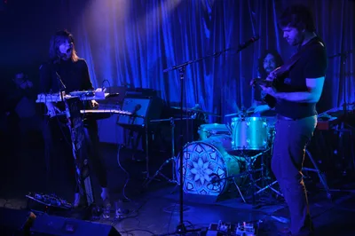 The experimental band Sun Blood Stories performs under moody blue stage lighting at Dante's in downtown Portland during Rx Fest. A keyboardist with shoulder-length hair commands the left side of the frame while a guitarist stands prominently on the right, with a drummer visible in the atmospheric shadows between them. The Boise-founded group, known for their opera-like sound and now based in Portland, creates a mesmerizing presence in the intimate venue's draped backdrop and professional lighting that captures the raw energy of their live performance.
