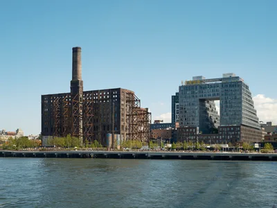 The skeletal remains of the historic Domino Sugar Factory stand sentinel against a cloudless blue sky, its weathered brick facade and exposed steel framework creating a dramatic contrast with the sleek glass towers of 325 Kent Avenue rising behind it. The East River flows in the foreground, reflecting the afternoon light as pedestrians stroll along the Williamsburg waterfront promenade. This striking juxtaposition captures Brooklyn's ongoing transformation from industrial past to contemporary urban landscape.