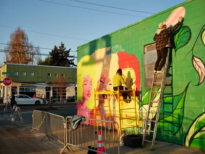 Multidisciplinary artist Vilatose works from a yellow scissor lift to paint the colorful portrait mural 'Jardín Cubana' depicting three female figures on a green cinderblock wall in Portland's Alberta Arts District. The golden hour light bathes the scene as the artist adds details to bold faces rendered in pink, yellow, and coral tones, while safety barriers protect the workspace below. The emerging artwork transforms the urban streetscape with its dynamic mix of portraiture and botanical elements.