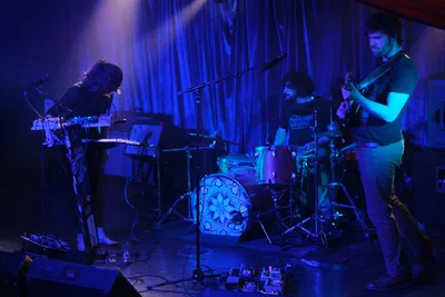 Boise experimental band Sun Blood Stories delivers an atmospheric performance at Rx Fest within the intimate confines of Dante's in downtown Portland. Deep purple and blue stage lighting bathes the three-piece band as they navigate their sonic landscape - the keyboardist hunched over synthesizers on the left, drummer maintaining rhythm behind a geometric kick drum design, and bassist anchored on the right. The moody illumination transforms the small venue into a nocturnal sanctuary where electronic textures merge with live instrumentation, creating the mesmerizing sound that has been likened to modern-day opera.