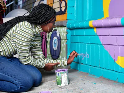 Naomi Likayi kneels on the sidewalk in Alberta Arts District, carefully painting her signature in purple letters across the vibrant turquoise and lavender geometric mural behind her. Her intricate box braids frame her concentrated expression as she works with a small brush, a paint can beside her on the concrete. The warm afternoon light illuminates her striped long-sleeve shirt and the bold street art that transforms this Northeast Portland corner into a canvas of community expression.