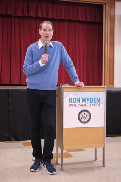 U.S. Senator Ron Wyden stands at a podium in the gymnasium of Robert Gray Middle School in Portland's Hillsdale neighborhood, addressing constituents during a town hall meeting. The senator, dressed casually in a blue sweater over a white collared shirt, grips a microphone while speaking against the backdrop of deep burgundy curtains that frame the school's performance space. The warm wooden podium bearing his official senatorial seal creates an intimate setting for this grassroots political engagement.