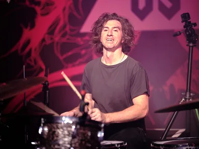 Julian Dorio, drummer for Band of Skulls, delivers an electrifying performance behind his kit at Manhattan's iconic Webster Hall during the band's 10 Year Anniversary Tour. Bathed in crimson stage lighting that transforms the intimate venue into a pulsing concert cathedral, Dorio's wild curls frame his concentrated expression as he grips his drumsticks mid-performance. The atmospheric lighting creates dramatic shadows across his simple black t-shirt while chrome cymbal surfaces catch and scatter the theatrical illumination throughout the storied East Village venue.