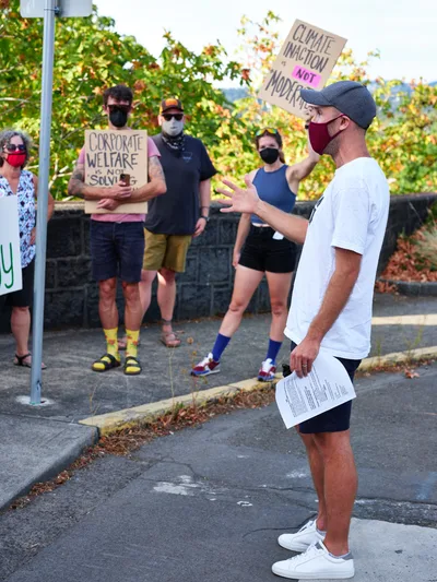 Peter Marks, wearing a red mask and baseball cap, gestures emphatically while addressing a small gathering of masked protesters outside Representative Kurt Schrader's Oregon City office. Behind him, activists hold handmade cardboard signs reading "CLIMATE INACTION NOT MODERATE" and "CORPORATE WELFARE IS NOT SOLVING," their colorful socks and casual attire contrasting with the serious political messaging. Golden autumn light filters through leafy trees, casting warm tones across the concrete sidewalk where this intimate act of civic engagement unfolds.