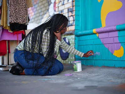 Naomi Likayi kneels on weathered pavement to sign her completed mural, her long braids cascading forward as she delicately applies her signature to the turquoise and purple brick wall. The scene captures an intimate moment of artistic completion in Portland's Alberta Arts District, where colorful murals transform urban spaces. Afternoon light illuminates the vibrant street art backdrop, creating a celebration of community creativity and cultural expression.