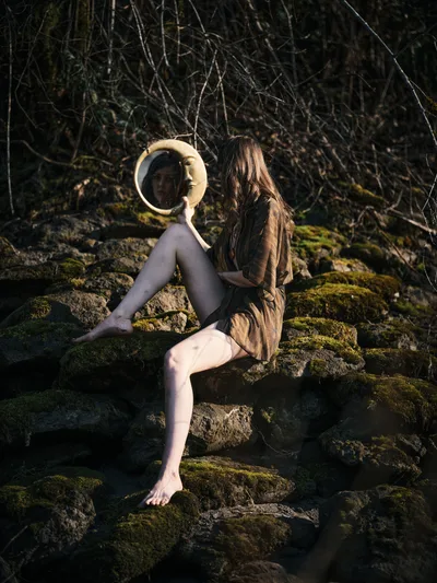 A contemplative figure sits barefoot on moss-covered rocks along a riverbed section of the Floodplain Trail in Multnomah County, Oregon, holding a circular mirror that captures their reflection. The subject wears an earth-toned jacket and dark leggings, positioned among weathered stones carpeted with vibrant green moss. Behind them, a dense tangle of bare winter branches creates a natural backdrop, while dappled sunlight filters through the forest canopy, casting warm highlights across the serene woodland scene.