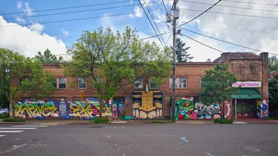 Vibrant street murals sprawl across the brick facade of the former Oregon Theater building on Southeast Division Street in Portland's Richmond neighborhood. The collaborative artwork, featuring bold geometric patterns, character portraits, and stylized lettering, creates a kaleidoscope of color against the weathered red brick under a dramatic cloudy sky. Power lines crisscross overhead while mature trees frame the scene, capturing the intersection of urban art and neighborhood history in this gentrifying corridor.