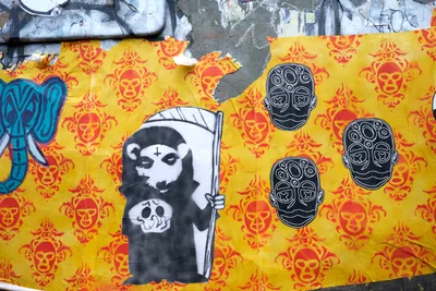 Weathered wheat paste murals blanket a concrete wall in Portland's Central Eastside, where mortality meets political commentary. A hooded Grim Reaper figure wielding a scythe dominates the left panel, juxtaposed against ornate sugar skull motifs scattered across vibrant yellow and red backgrounds. Three pristine white skulls with geometric patterns float like spectral guardians, their clean lines contrasting sharply with the deteriorating urban canvas beneath layers of peeling paint and exposed concrete.