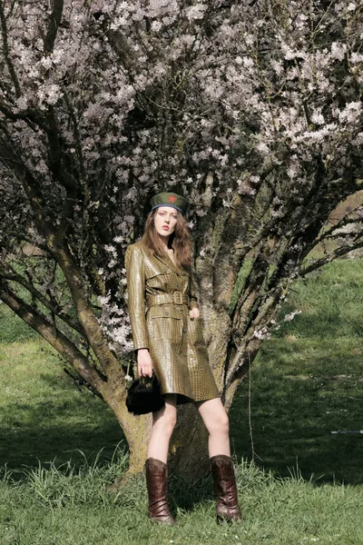 Model Rhianna Atwood poses beneath the delicate pink sakura blooms in Portland's Mount Tabor Park, wearing a military-inspired olive leather trench coat and burgundy boots. The spring light filters through the flowering branches, creating a dreamy hanami atmosphere against the verdant Oregon hillside. Her playful expression and vintage-style beret add whimsy to this seasonal editorial moment.