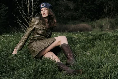 Model Rhianna Atwood reclines gracefully in tall grass wearing a structured olive military-inspired coat and navy beret, captured during golden hour at Mount Tabor Park in Portland, Oregon. The moody lighting creates dramatic shadows across the natural meadow setting, while bare winter trees frame the background. Her contemplative expression and relaxed pose against the earthy terrain evoke a sense of quiet rebellion and seasonal transition.